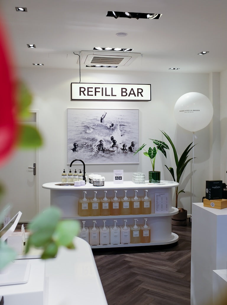 Inside the Marie-Stella-Maris boutique in Maastricht: glowing ‘Refill Bar’ sign above a curved white counter stocked with refill dispensers, a large black-and-white water photograph, potted plants, and dark herringbone wood floors.
