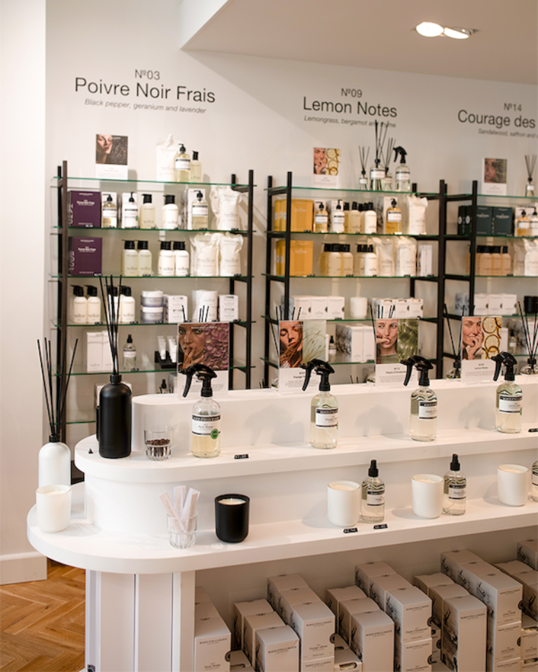 Inside the Marie-Stella-Maris boutique in Rotterdam: black metal shelves of body care and home fragrance organized by scent (Poivre Noir Frais, Lemon Notes, Courage des Bois), with tester sprays and candles on a curved white counter over herringbone flooring.