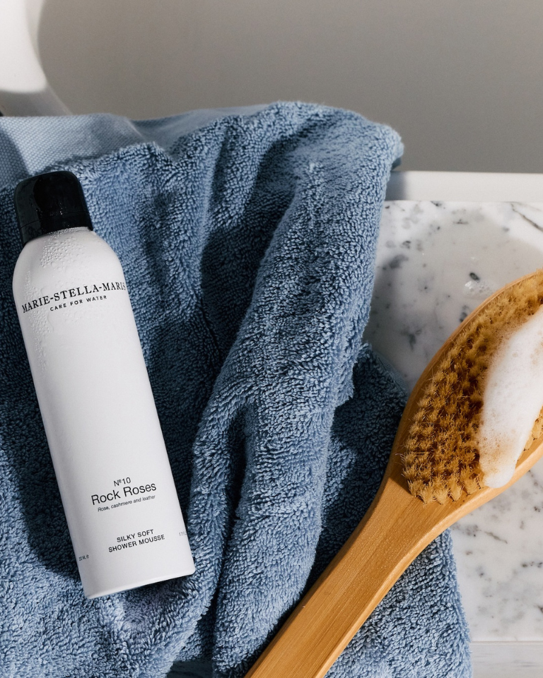 Marie-Stella-Maris Silky Soft Shower Mousse No.10 Rock Roses placed beside a soft blue towel and wooden body brush, creating a calm and luxurious bathroom setting.