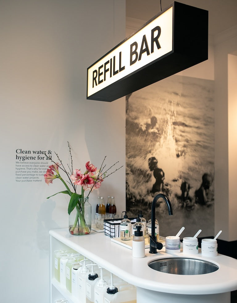 Inside the Marie-Stella-Maris boutique in Utrecht: glowing ‘Refill Bar’ lightbox over a curved white counter with a sink, refill pumps and jars, fresh flowers, and a large black-and-white water photograph with ‘Clean water & hygiene for all’ text on the wall.