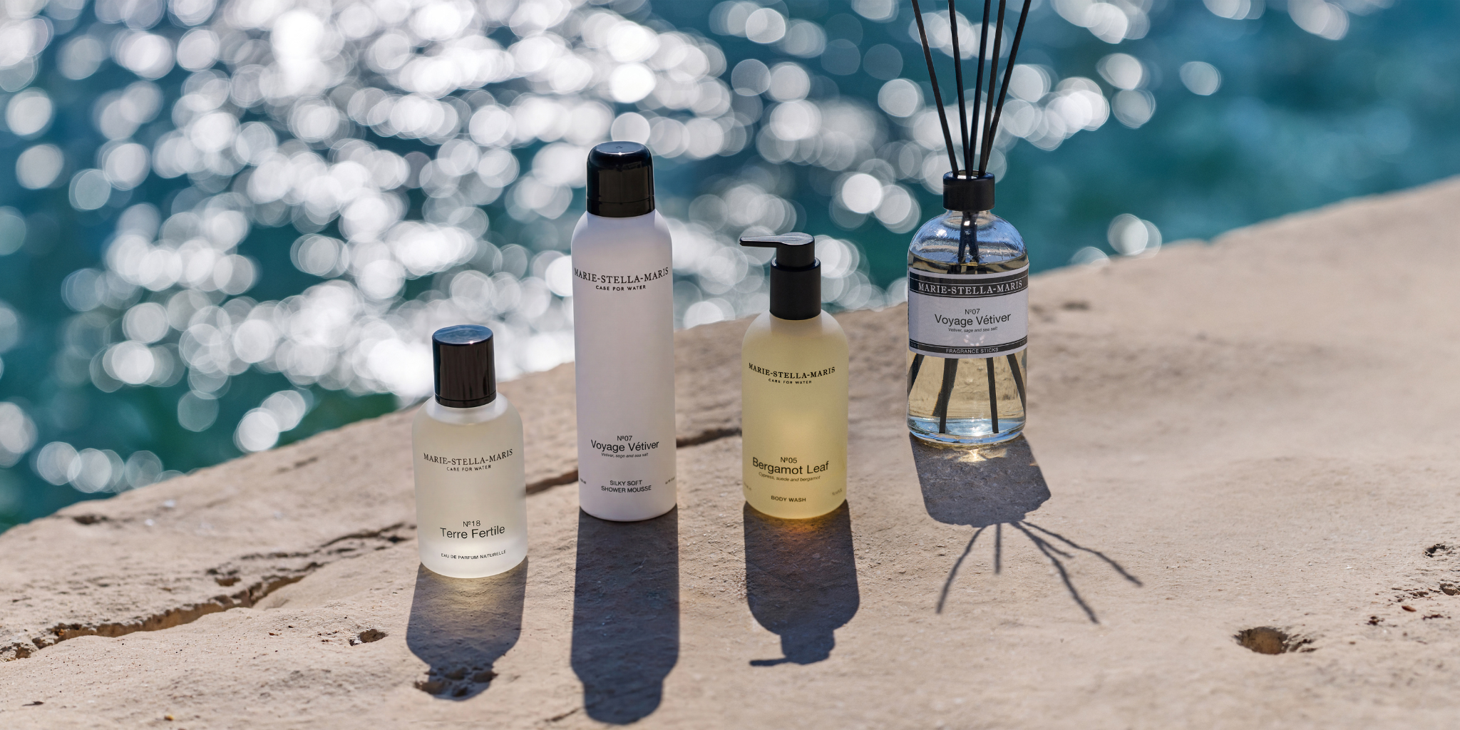 Marie-Stella-Maris Father’s Day selection featuring No.18 Terre Fertile eau de parfum naturelle, No.07 Voyage Vétiver Silky Soft Shower Mousse, No.05 Bergamot Leaf body wash, and No.07 Voyage Vétiver fragrance sticks, displayed on a sunlit stone surface by the sea.