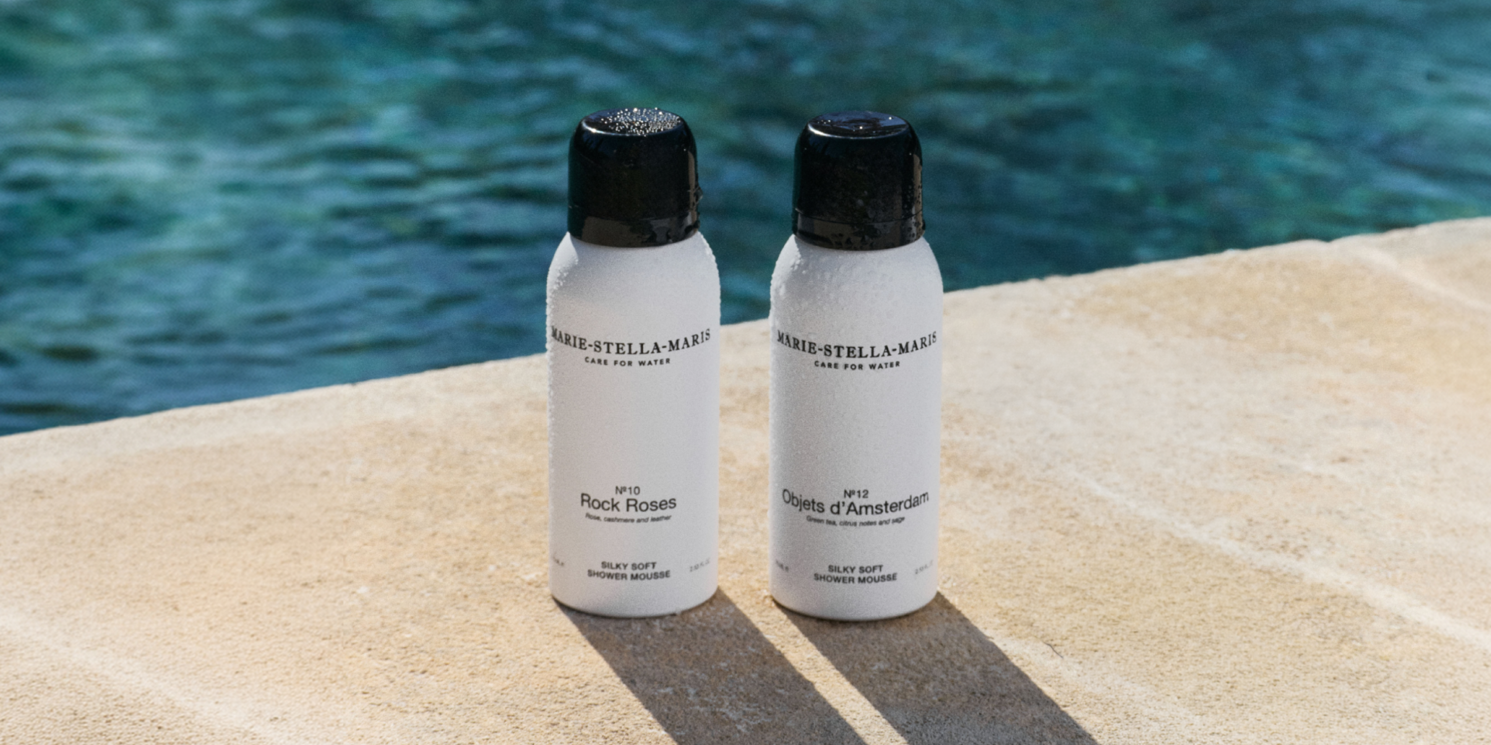 Marie-Stella-Maris 75 ml travel-size Silky Soft Shower Mousses in No.10 Rock Roses and No.12 Objets d’Amsterdam, displayed on a sunlit stone surface by the water with cooling droplets on the bottles, highlighting the summer travel essentials collection.