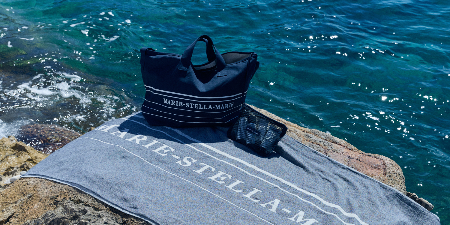 Marie-Stella-Maris summer essentials displayed on a rocky shoreline, featuring the limited edition gift with purchase collection: a navy blue summer tote bag, matching towel, and black mesh pouch, set against sparkling blue sea water.