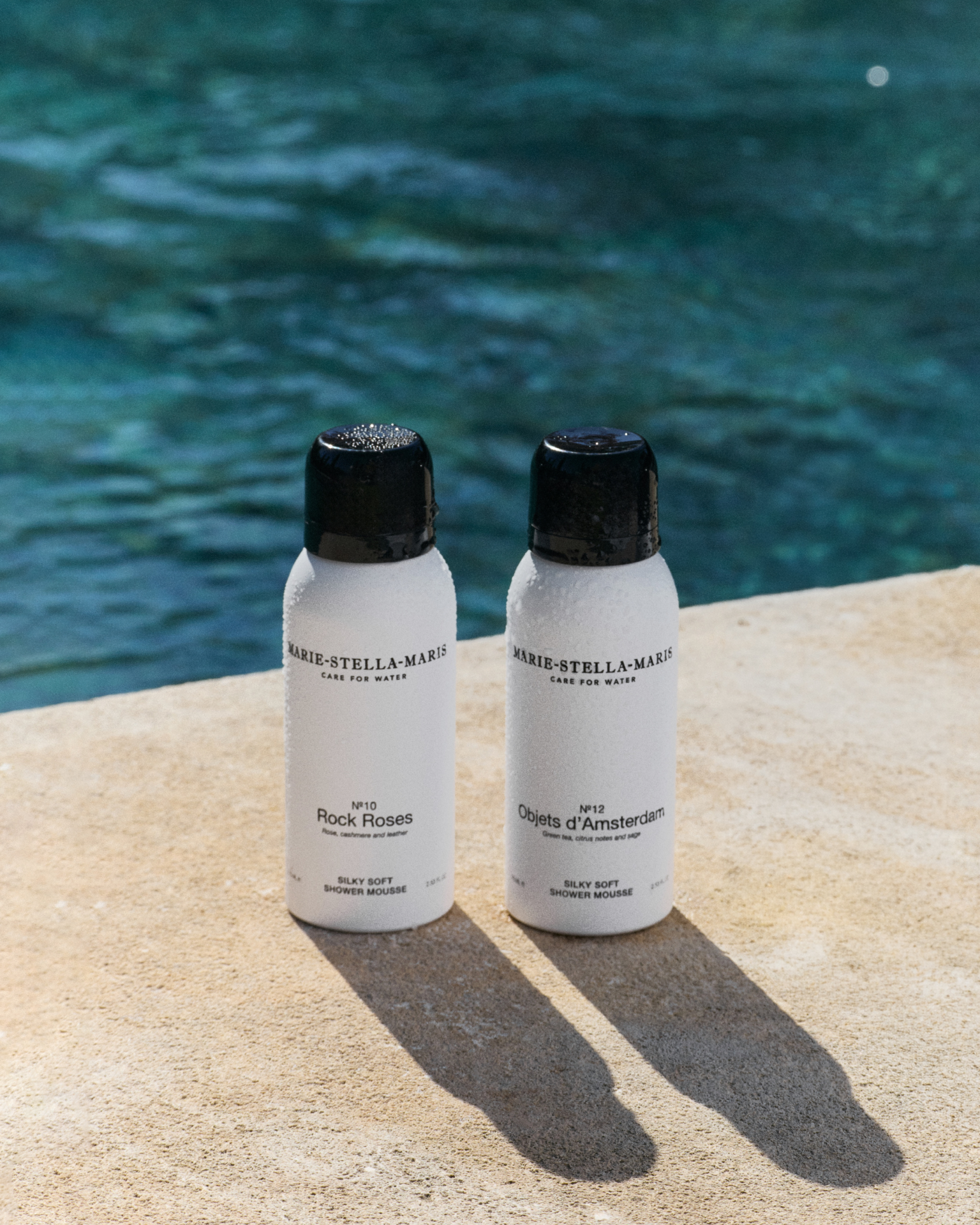 Marie-Stella-Maris 75 ml travel-size Silky Soft Shower Mousses in No.10 Rock Roses and No.12 Objets d’Amsterdam, displayed on a sunlit stone surface by the water with cooling droplets on the bottles, highlighting the summer travel essentials collection.