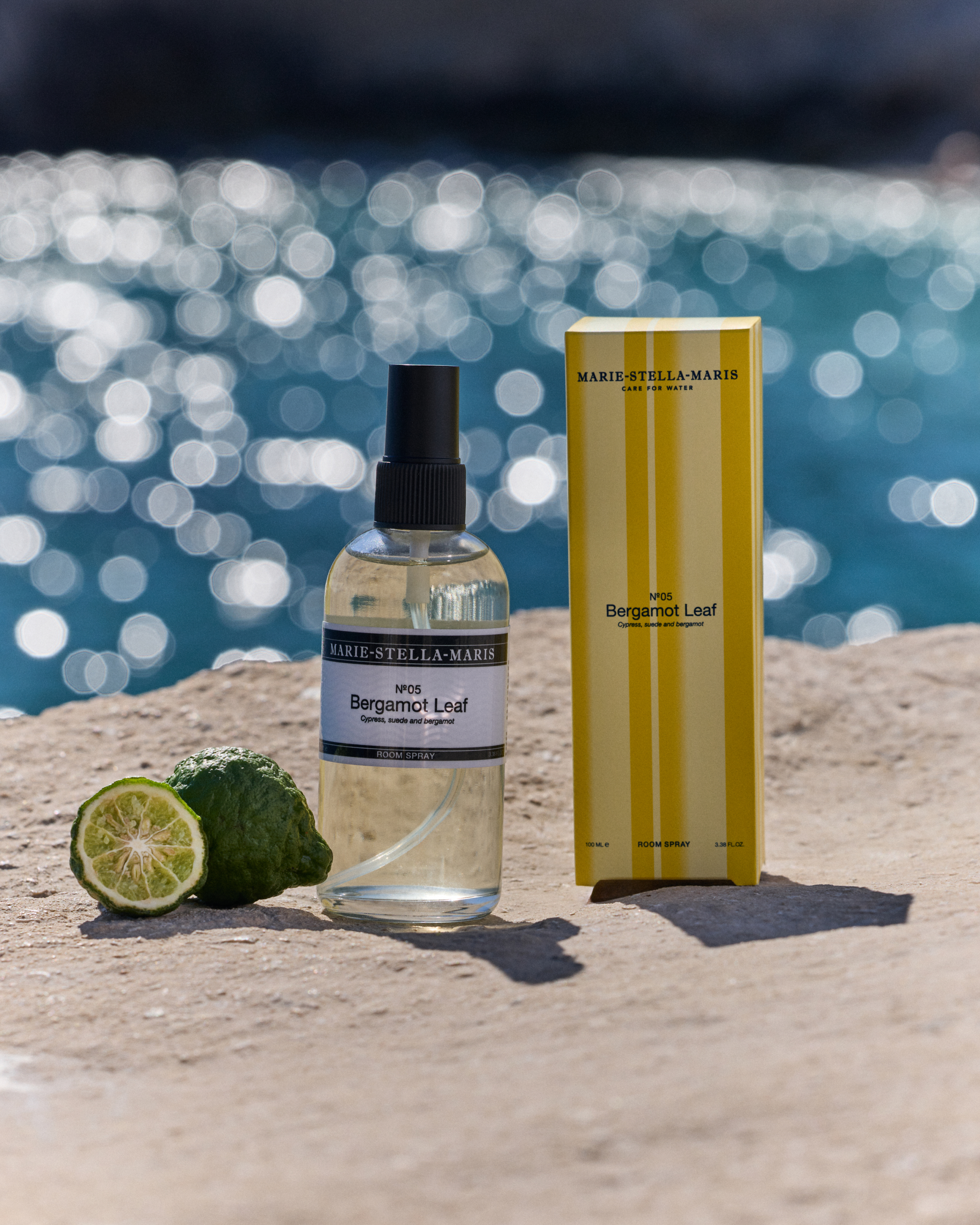Marie-Stella-Maris No.05 Bergamot Leaf limited edition summer scent room spray 100 ml with matching yellow-striped packaging, displayed on a sunlit stone surface by the sea with fresh bergamot and sparkling water in the background.