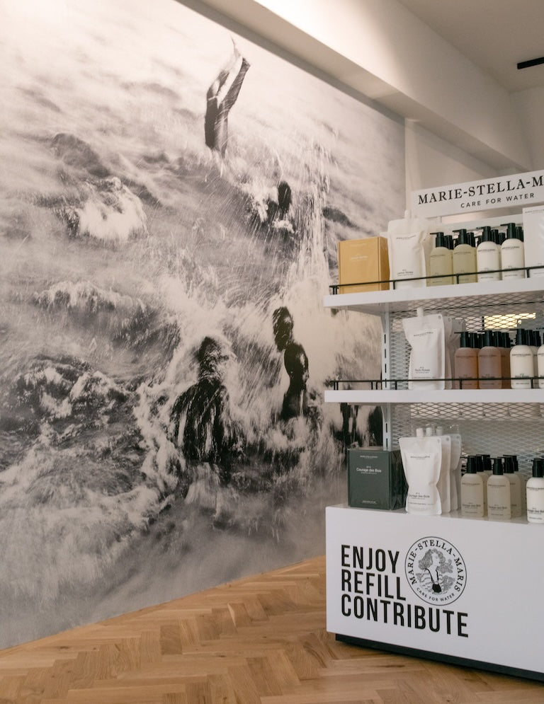 Inside the Marie-Stella-Maris Amsterdam Zuid boutique: floor-to-ceiling black-and-white water mural beside shelves of body care and refills with ‘Enjoy Refill Contribute’ branding.