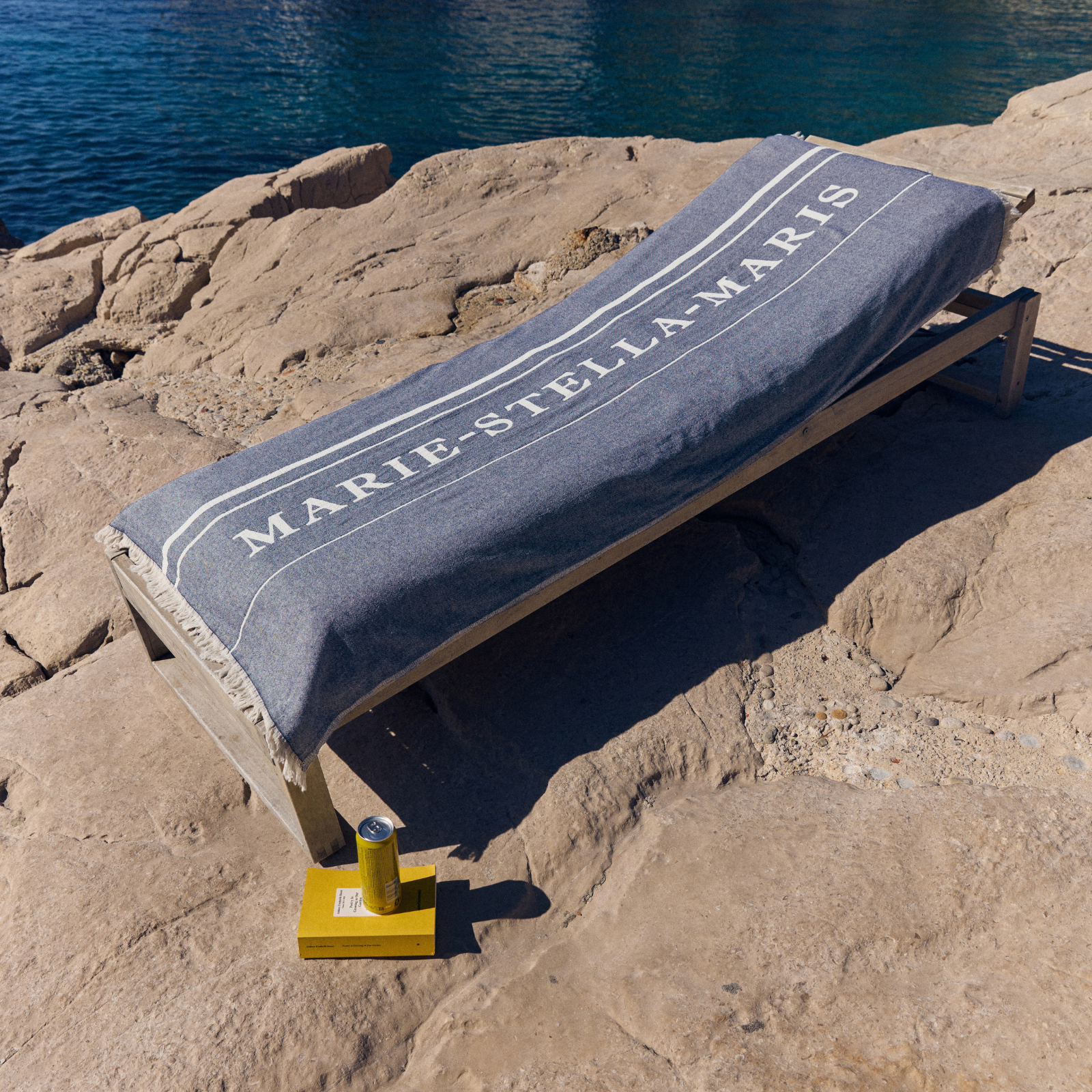 Marie-Stella-Maris towel draped over a sunbed on a rocky shoreline, part of the limited edition summer 2026 gift with purchase collection, with the sea and coastal landscape in the background, capturing a relaxed summer atmosphere.