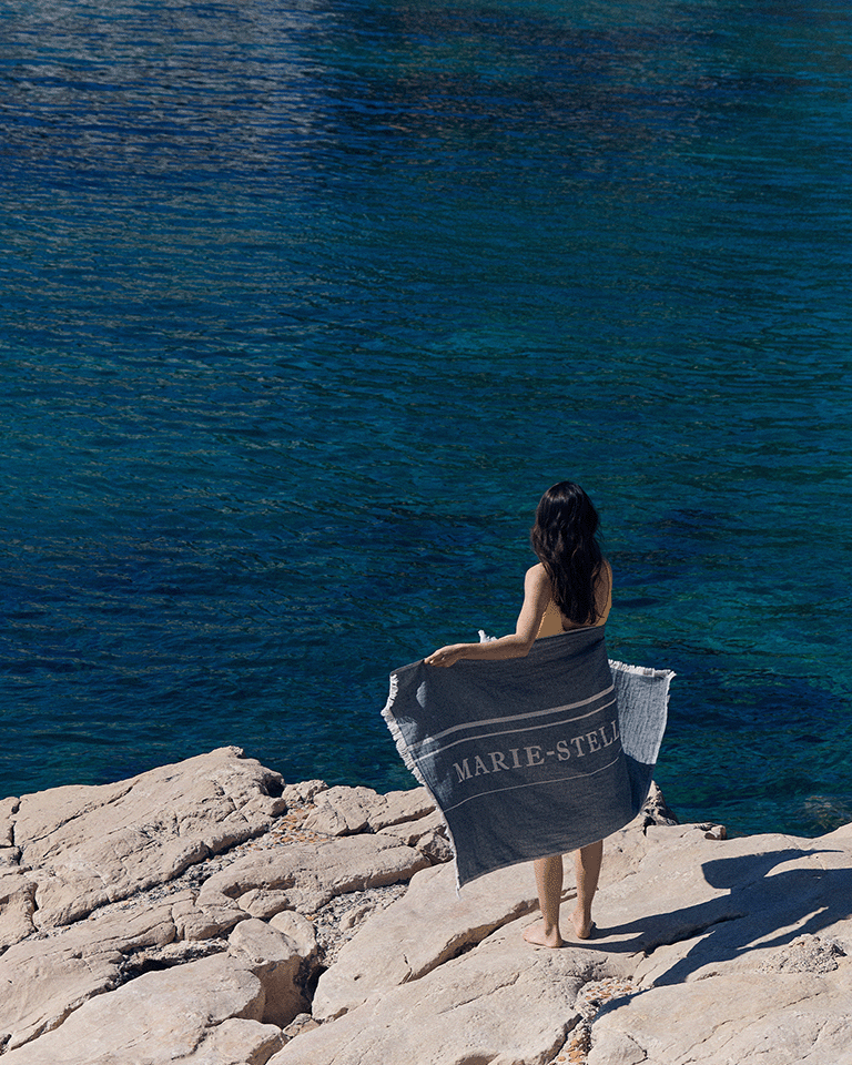 Person standing on a rocky shoreline wrapped in a Marie-Stella-Maris towel, part of the limited edition gift with purchase collection, overlooking clear blue sea.