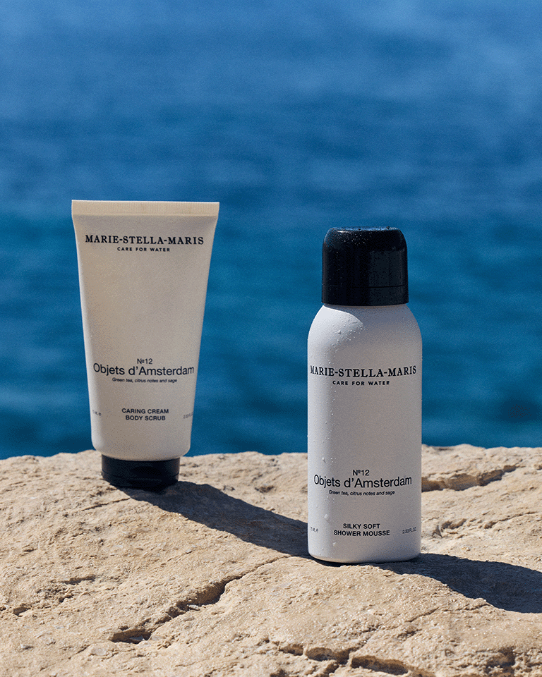 Marie-Stella-Maris Objets d’Amsterdam Caring Cream Body Scrub and Silky Soft Shower Mousse displayed on sunlit coastal rocks with a deep blue sea in the background. The white packaging contrasts with the natural stone and ocean setting.