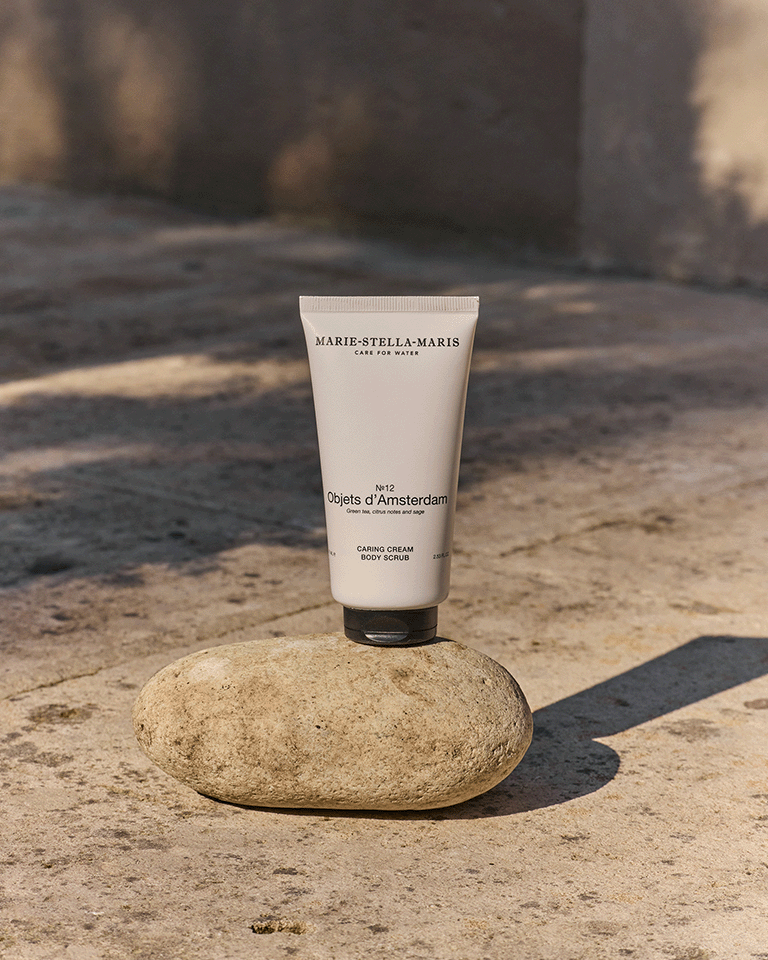 Marie-Stella-Maris Objets d’Amsterdam Caring Cream Body Scrub standing upright on a smooth stone in warm natural light. The product is showcased against a softly blurred outdoor backdrop, emphasizing its minimalist design and premium skincare appeal.