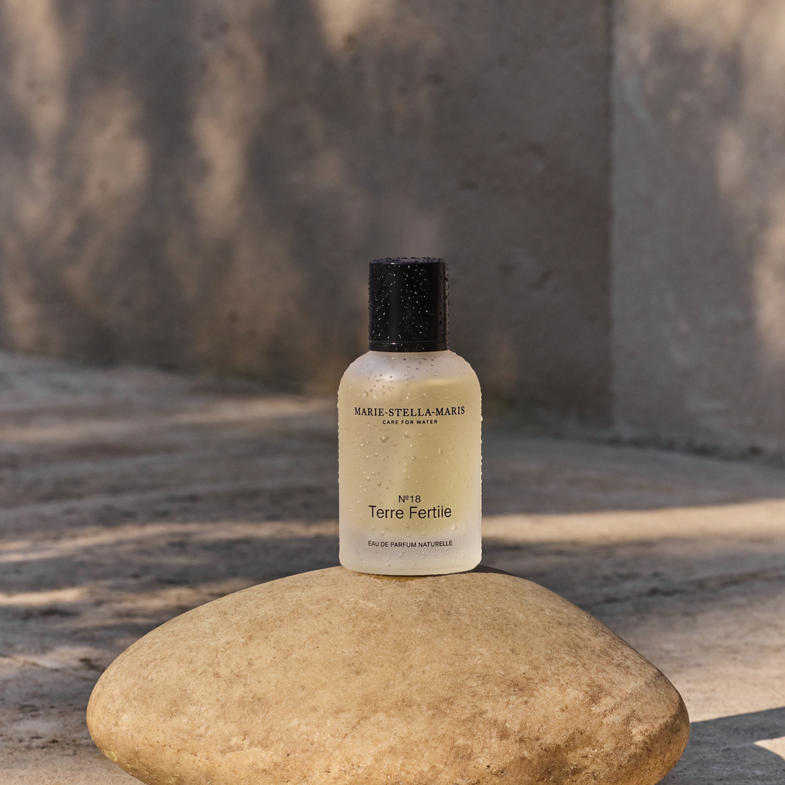 Marie-Stella-Maris No.18 Terre Fertile eau de parfum naturelle, displayed on a smooth stone in a sunlit outdoor setting with water droplets on the bottle, highlighting the fragrance in a warm and refined atmosphere.