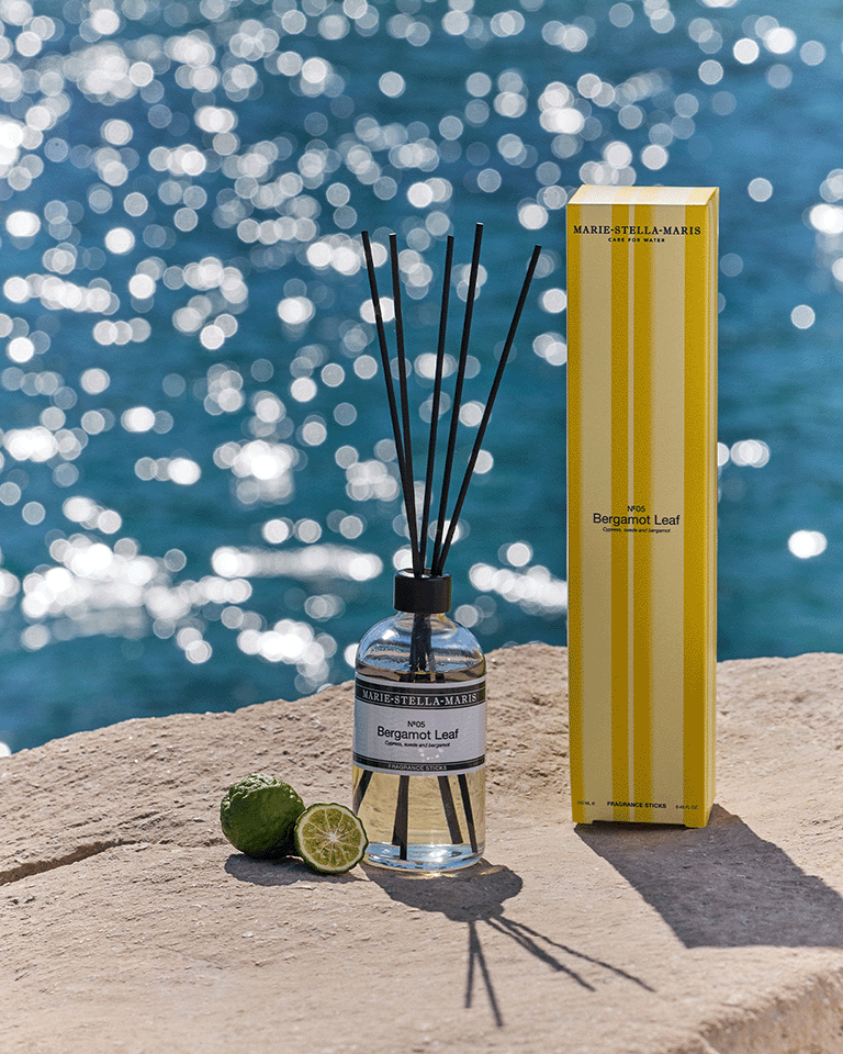 Marie-Stella-Maris Bergamot Leaf fragrance diffuser with black reeds, displayed beside its yellow-striped box on a sunlit stone surface, with fresh limes and sparkling blue water in the background.