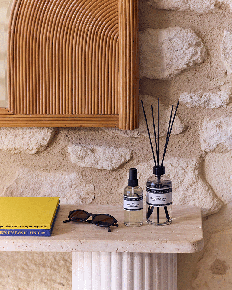 Marie-Stella-Maris fragrance sticks 250 ml and Room Spray 100 ml in the No.05 Bergamot Leaf scent, displayed on a stone table against a textured wall, with a wooden framed mirror, sunglasses, and stacked books nearby.