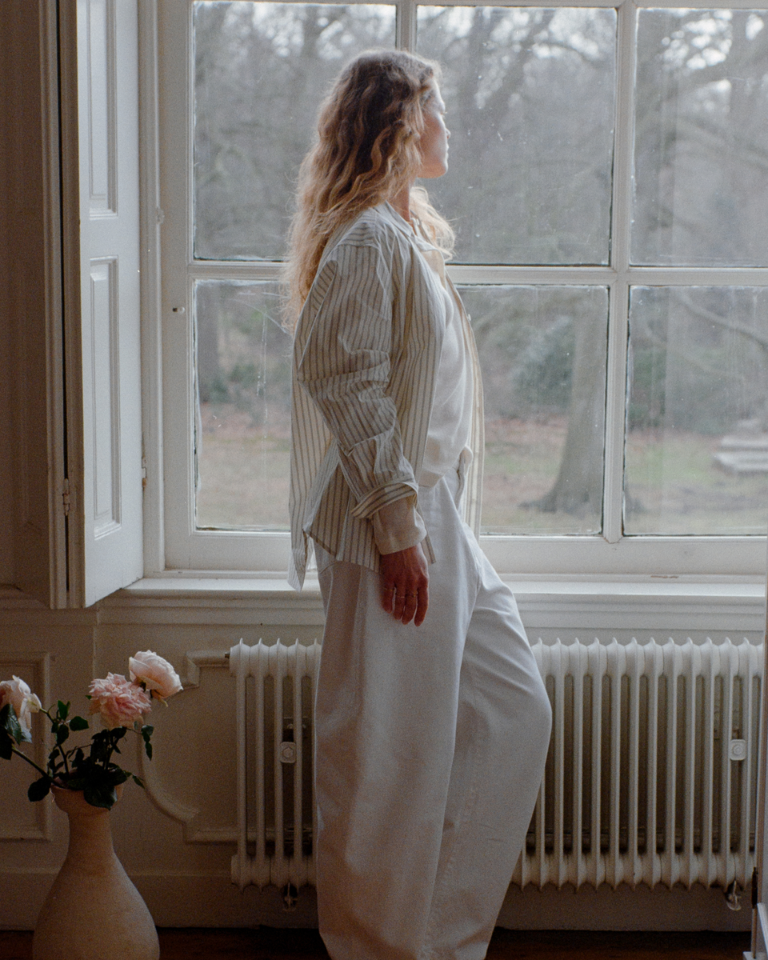 Person standing by a window in a calm indoor setting, wearing light, relaxed clothing, with soft natural light and a vase of pink roses nearby, creating a serene atmosphere for the Marie-Stella-Maris Mother’s Day campaign.