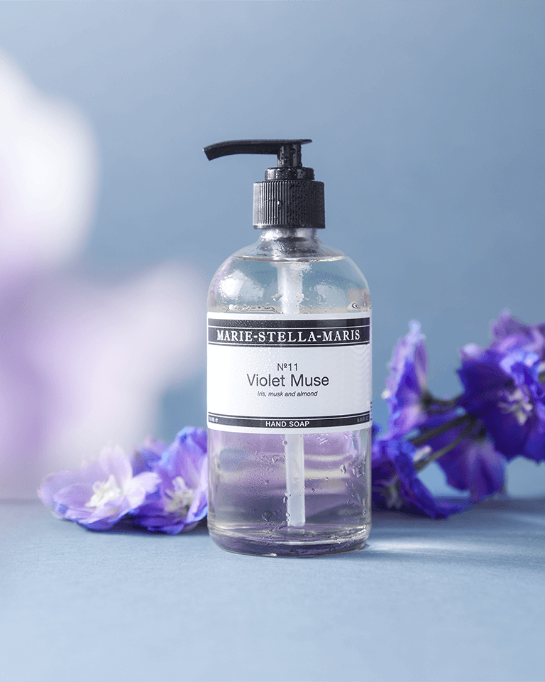 Marie-Stella-Maris Hand Soap No.11 Violet Muse 250 ml displayed with purple flowers, featuring a transparent bottle with a black pump and notes of iris, musk and almond.