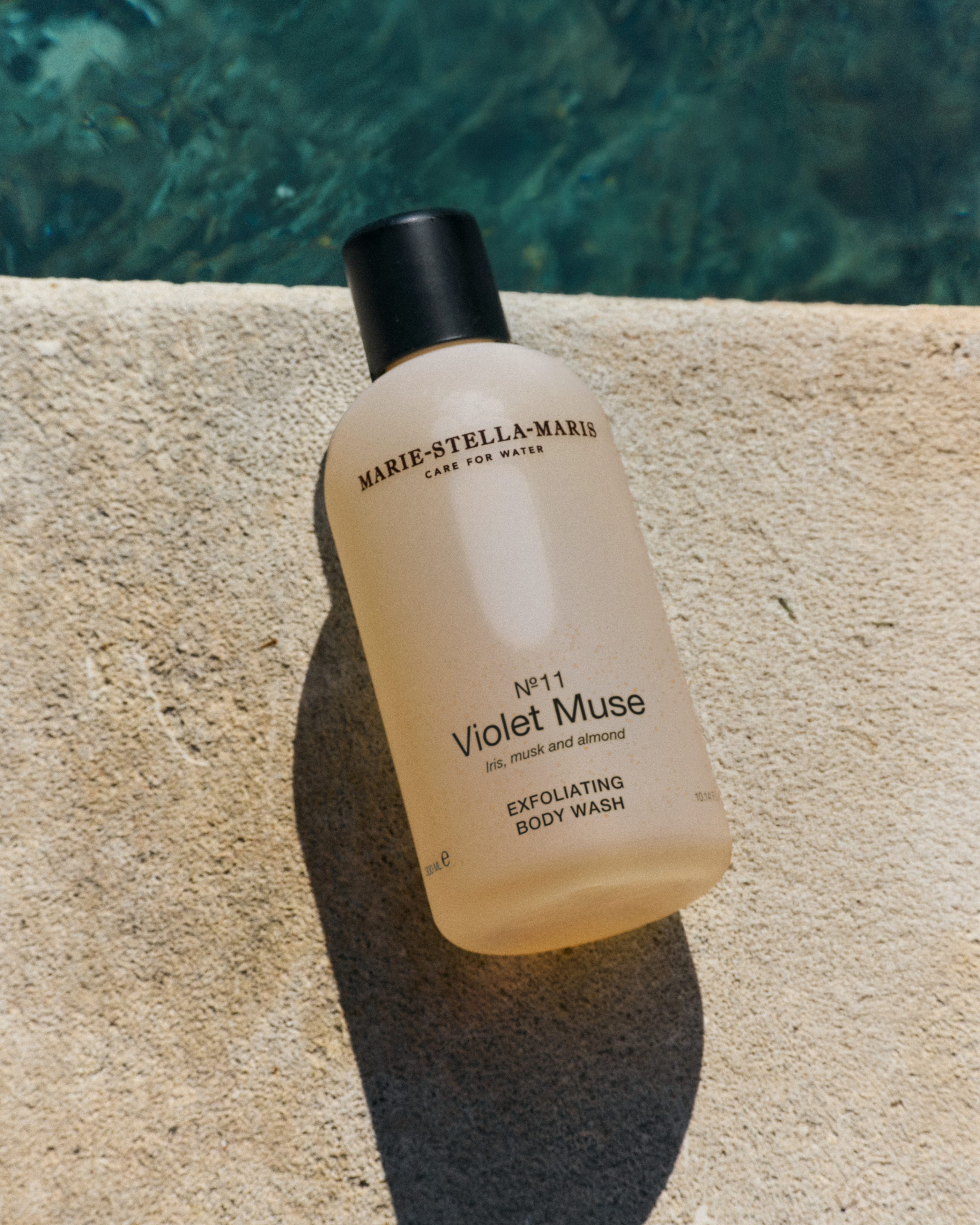 Marie-Stella-Maris No.11 Violet Muse exfoliating body wash 300 ml, displayed on a sunlit stone surface beside clear blue water, highlighting fragrance notes of iris, musk, and almond in a fresh summer setting.