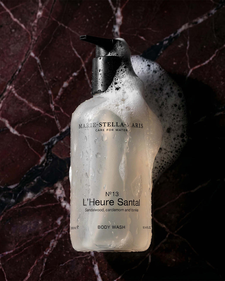 Close-up of Marie-Stella-Maris No.13 L'Heure Santal Body Wash (300 ml) covered in water droplets and rich foam against a dark marble surface. The image highlights the body wash's luxurious texture and its signature fragrance with notes of sandalwood, cardamom and tonka.