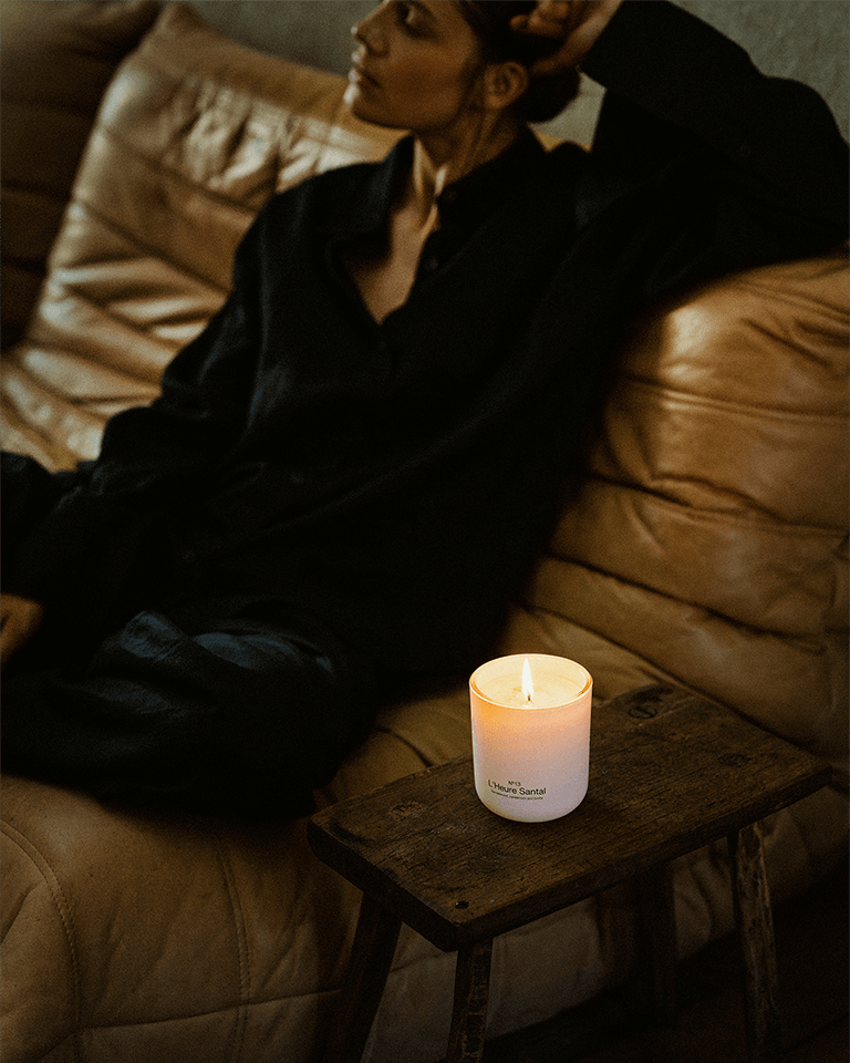 Marie-Stella-Maris No.13 L'Heure Santal Scented Candle burning on a rustic wooden stool beside a leather sofa, creating a warm and inviting atmosphere. A person relaxes in the background, reflecting the comforting character of the fragrance with notes of sandalwood, cardamom and tonka.
