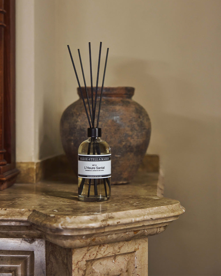 Marie-Stella-Maris No.13 L'Heure Santal Fragrance Sticks (250 ml) displayed on a marble mantelpiece beside a rustic ceramic vase. The black fragrance reeds and warm interior setting reflect the elegant character of the fragrance with notes of sandalwood, cardamom and tonka.