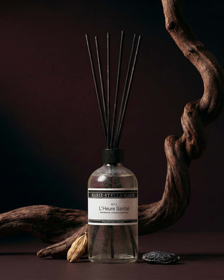Marie-Stella-Maris No.13 L'Heure Santal Fragrance Sticks (250 ml) displayed with black fragrance reeds against a dark, elegant backdrop. Twisted wood, cardamom pods and textured stones surround the diffuser, reflecting the fragrance's warm notes of sandalwood, cardamom and tonka.