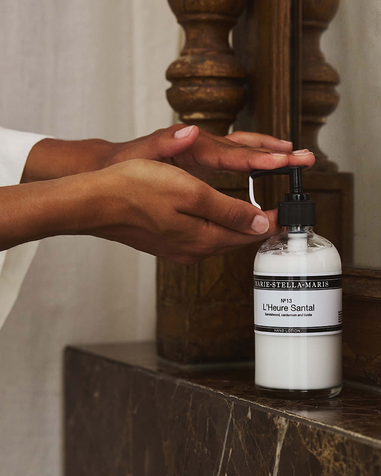 Hands dispensing Marie-Stella-Maris No.13 L'Heure Santal Hand Lotion from a pump bottle placed on a marble countertop. The warm interior setting complements the fragrance's refined notes of sandalwood, cardamom and tonka.
