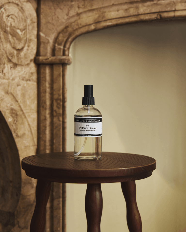 Marie-Stella-Maris No.13 L'Heure Santal Room Spray (250 ml) displayed on a dark wooden side table in front of an elegant stone fireplace. The warm interior setting complements the room spray's refined fragrance with notes of sandalwood, cardamom and tonka.