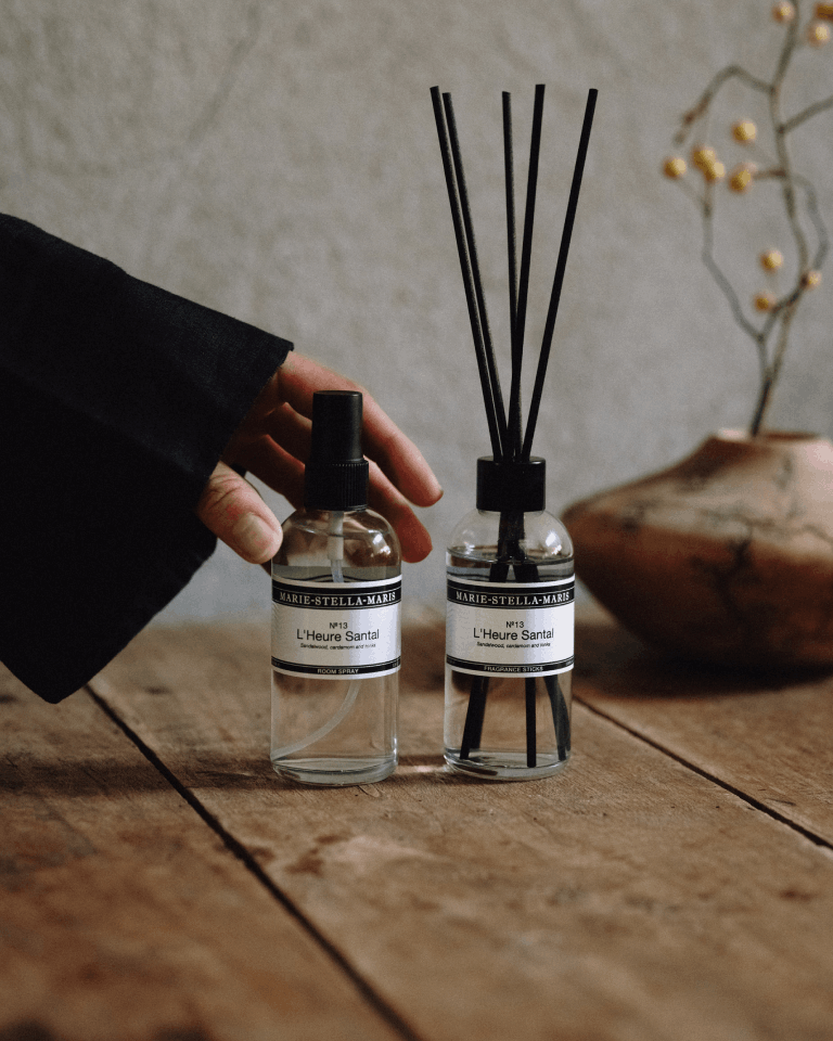 Marie-Stella-Maris No.13 L'Heure Santal Room Spray (100 ml) and Fragrance Sticks displayed together on a rustic wooden table. A hand reaches towards the room spray, while the diffuser with black fragrance reeds creates a warm and inviting atmosphere inspired by notes of sandalwood, cardamom and tonka.