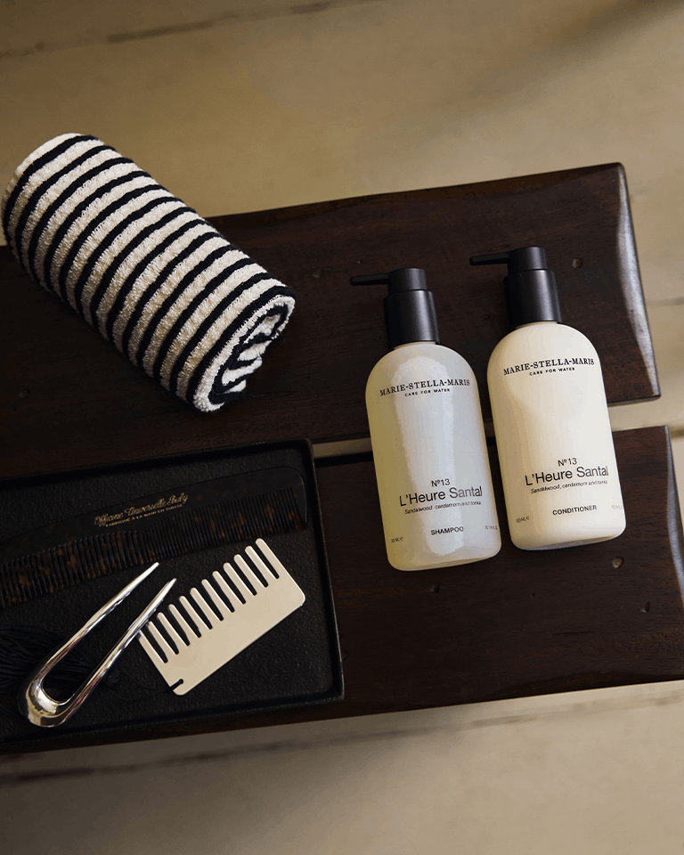 Marie-Stella-Maris No.13 L'Heure Santal Shampoo and Conditioner (300 ml) displayed on a dark wooden bench alongside a striped towel, comb and hair accessories. The warm bathroom setting highlights the hair care duo and the signature fragrance with notes of sandalwood, cardamom and tonka.
