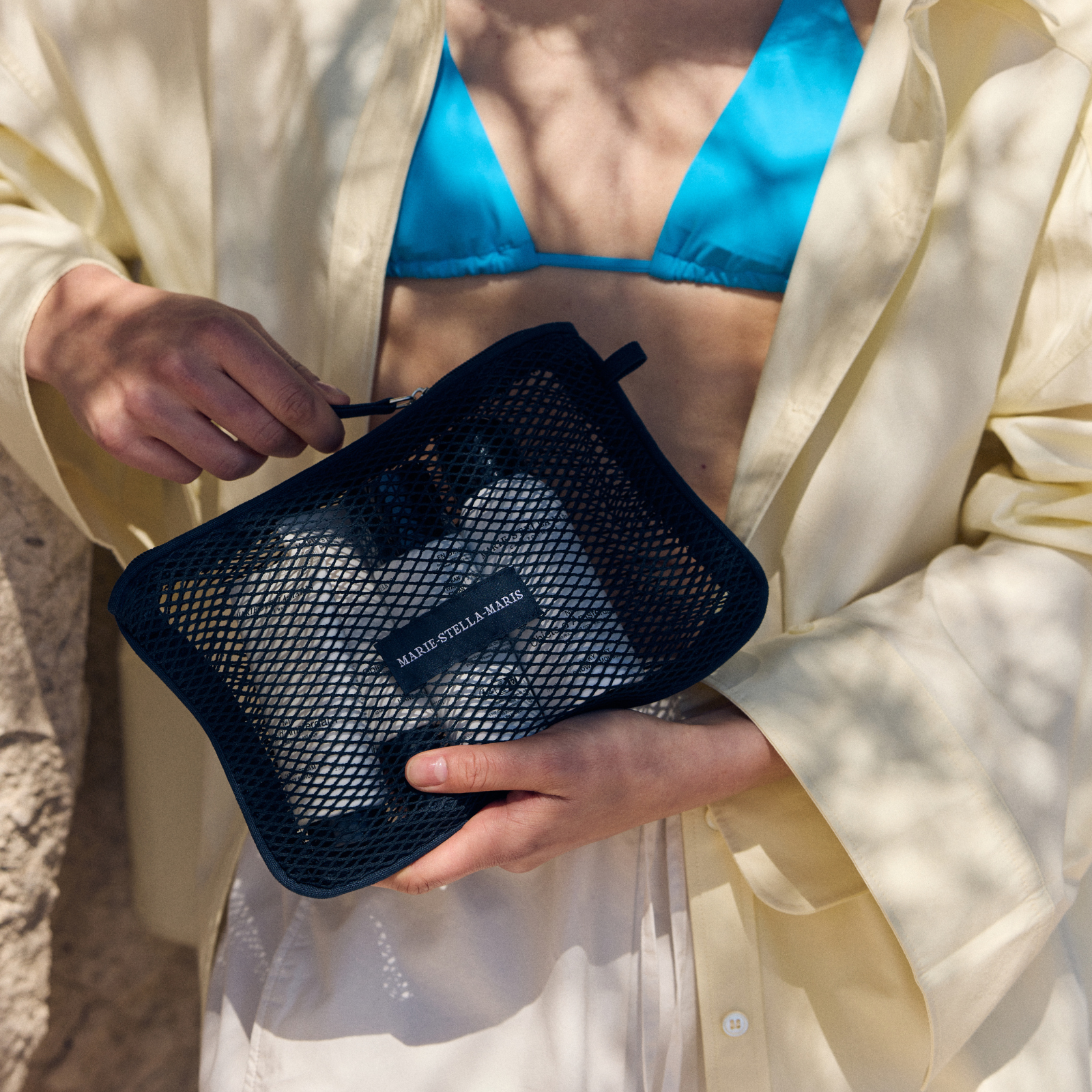 Person wearing a blue bikini top and light shirt holding a Marie-Stella-Maris black mesh pouch, part of the limited edition summer 2026 gift with purchase collection, with travel-size body care essentials visible inside and soft sunlight casting shadows across the scene.