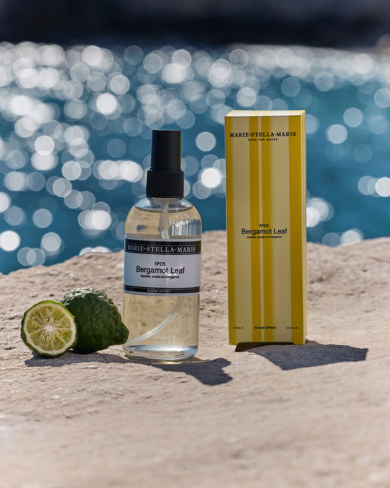 Marie-Stella-Maris No.05 Bergamot Leaf limited edition summer scent room spray 100 ml with matching yellow-striped packaging, displayed on a sunlit stone surface by the sea with fresh bergamot fruit and sparkling water in the background.