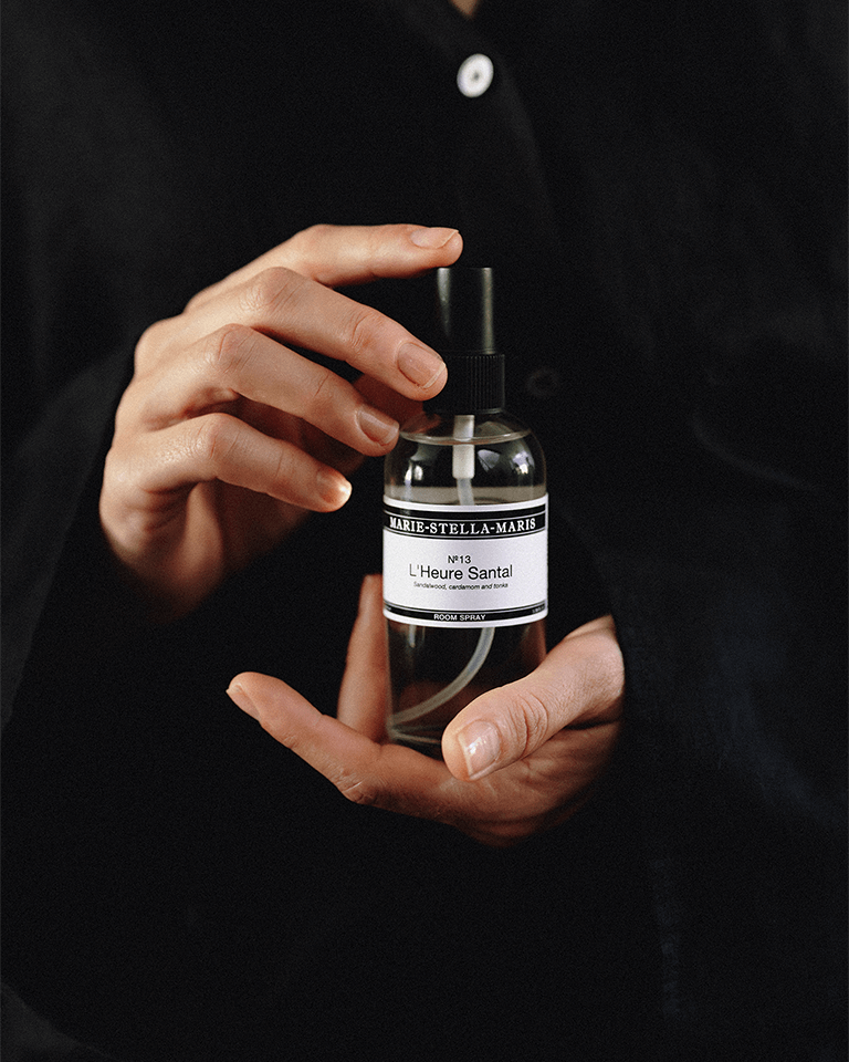 Close-up of hands holding a Marie-Stella-Maris No.13 L'Heure Santal Room Spray (100 ml). The minimalist spray bottle is set against a dark background, highlighting the elegant design and the fragrance's notes of sandalwood, cardamom and tonka.