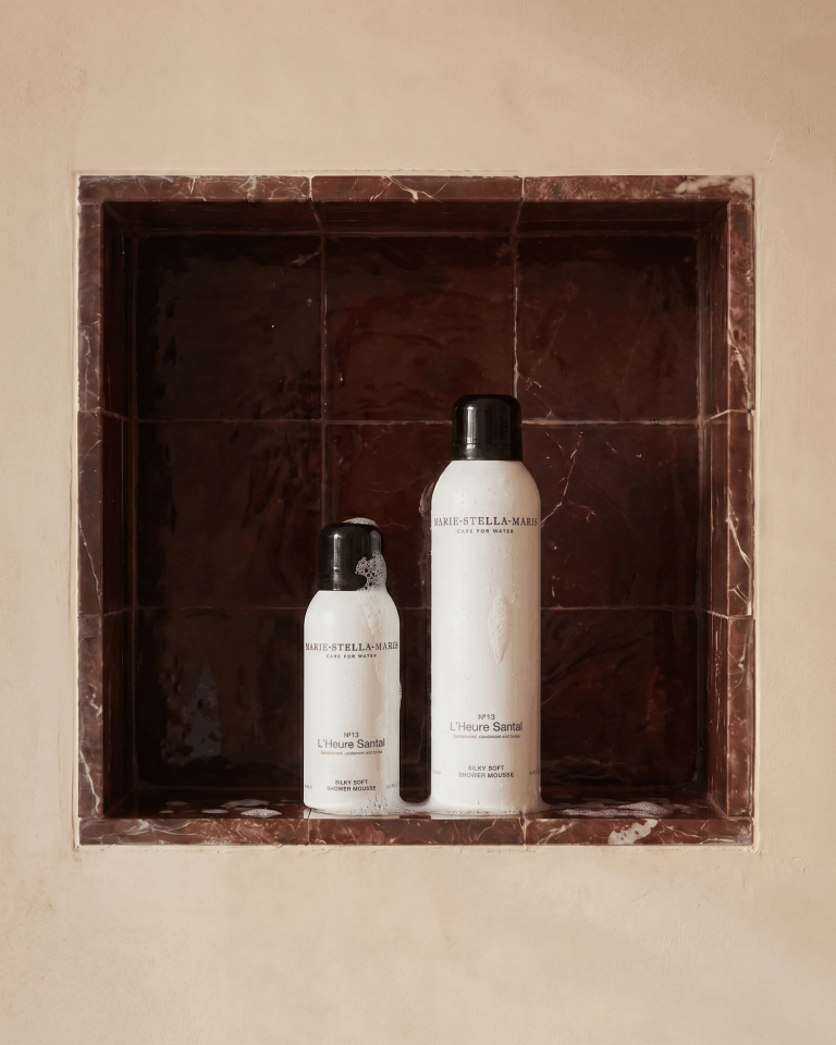 Marie-Stella-Maris No.13 L'Heure Santal Silky Soft Shower Mousse in 75 ml and 300 ml sizes displayed together in a tiled shower niche. The white bottles with black caps are accented with soft foam, highlighting the luxurious shower experience and the fragrance's notes of sandalwood, cardamom and tonka.
