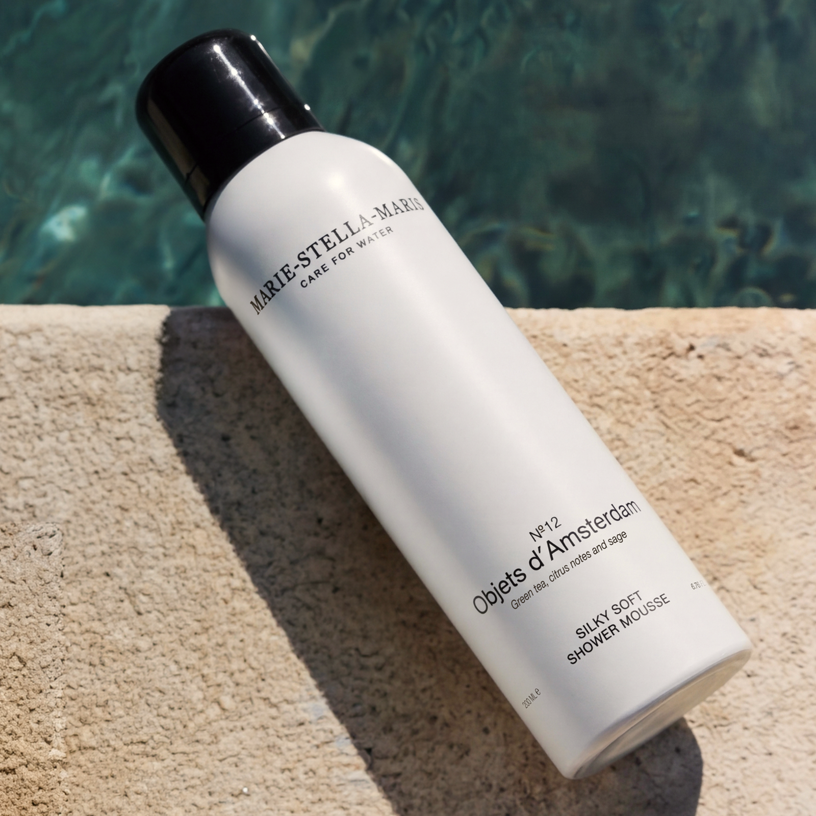 Marie-Stella-Maris No.12 Objets d’Amsterdam Silky Soft Shower Mousse 200 ml, displayed on a sunlit stone surface beside clear blue water, highlighting fragrance notes of green tea, citrus notes, and sage in a fresh summer setting.