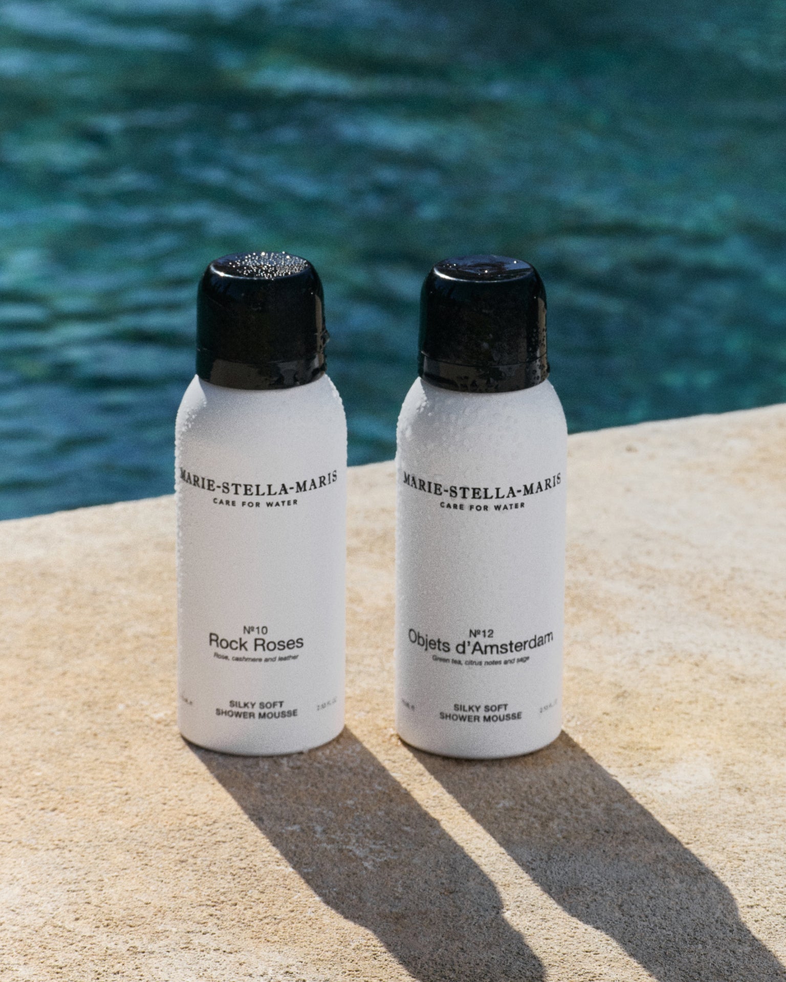 Marie-Stella-Maris 75 ml travel-size Silky Soft Shower Mousse bottles in No.10 Rock Roses, No.12 Objets d’Amsterdam, and No.13 L’Heure Santal, placed on a sunlit stone surface by the sea with water droplets on the bottles and ocean in the background.