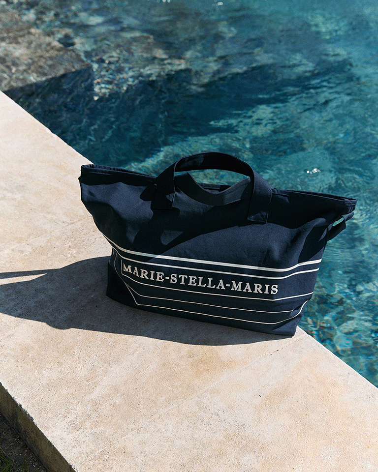 Marie-Stella-Maris limited edition summer 2026 navy blue tote bag with white stripe detailing and logo, placed on a sunlit poolside edge beside clear blue water.
