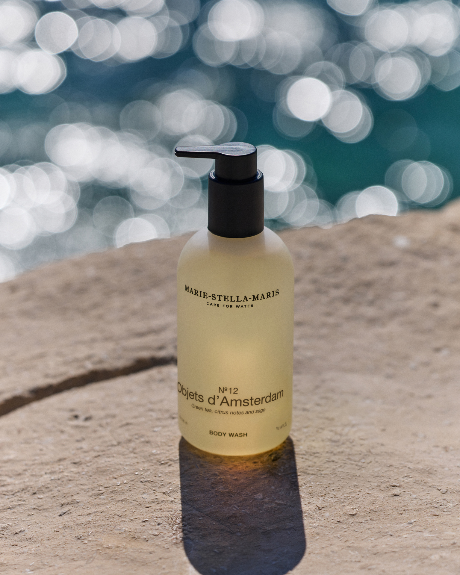 Marie-Stella-Maris No.12 Objets d’Amsterdam body wash 300 ml, featured as a May gift with purchase, displayed on a sunlit stone surface by the sea with sparkling water in the background.