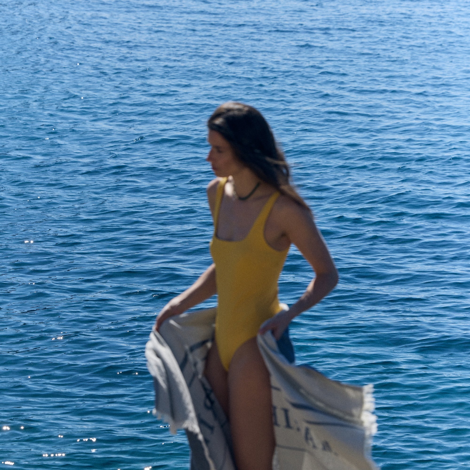 Person by the sea wearing a yellow swimsuit and holding a towel, capturing a relaxed, sunlit summer moment aligned with the Marie-Stella-Maris summer campaign.