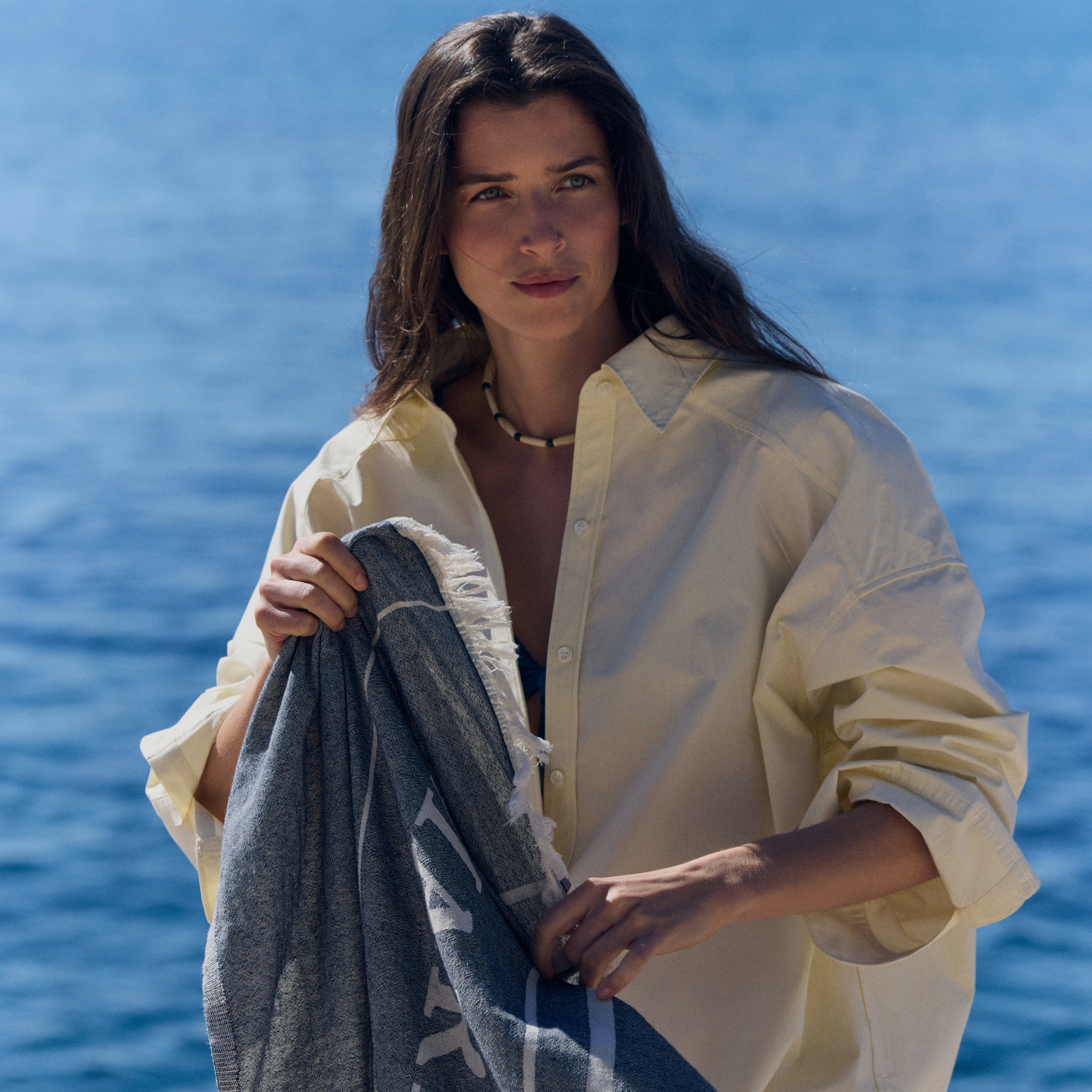Person standing by the sea wearing a light shirt and holding a Marie-Stella-Maris towel, part of the limited edition gift with purchase collection, with sparkling blue water in the background, capturing the relaxed spirit of the Marie-Stella-Maris summer campaign.