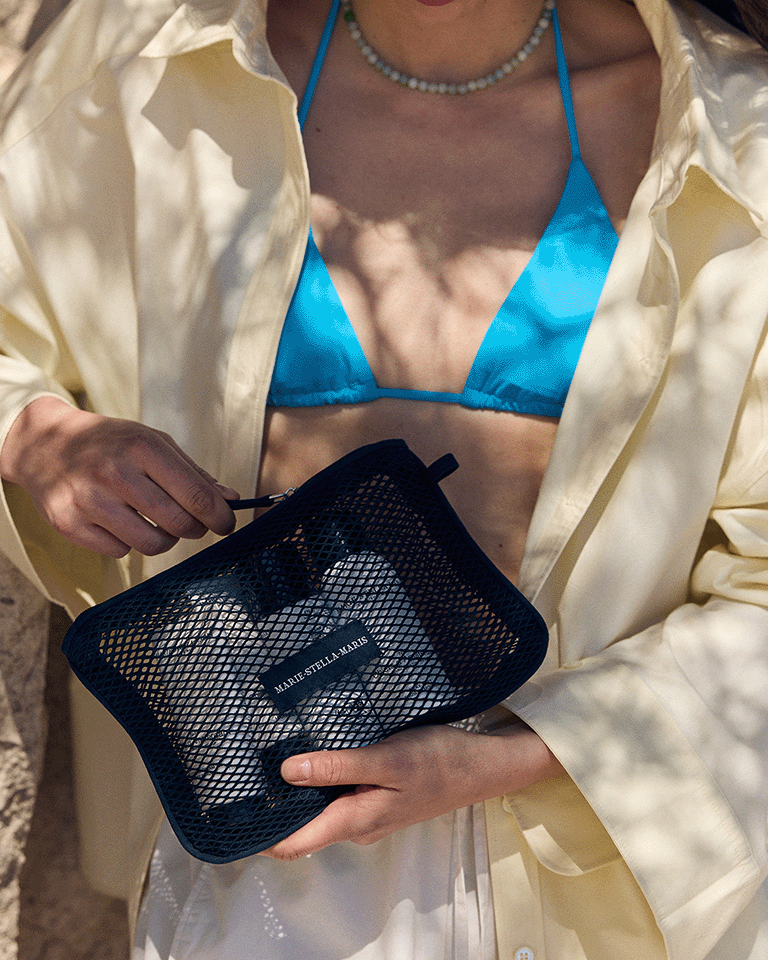Person wearing a blue bikini top and light shirt holding a Marie-Stella-Maris black mesh pouch, with sunlight casting soft shadows across the scene.