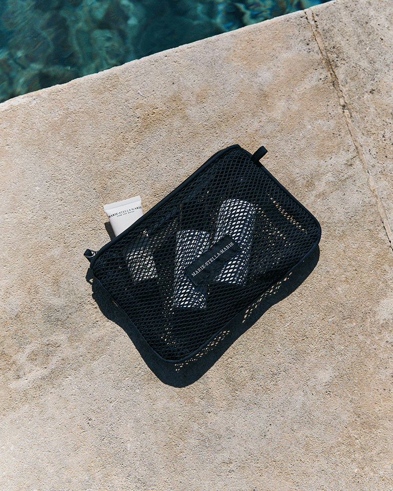 Marie-Stella-Maris beach setup with a blue towel laid on rocky shore, a matching tote bag and black mesh pouch placed beside it, overlooking clear sparkling sea water.