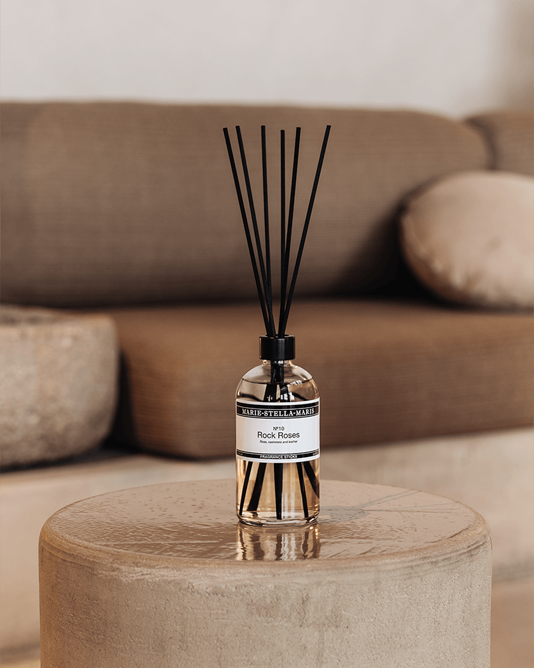 Marie-Stella-Maris Fragrance Sticks No.10 Rock Roses 250 ml in a clear glass bottle with black reeds, placed on a textured round table in a warm, minimalist living room setting with a soft sofa in the background.