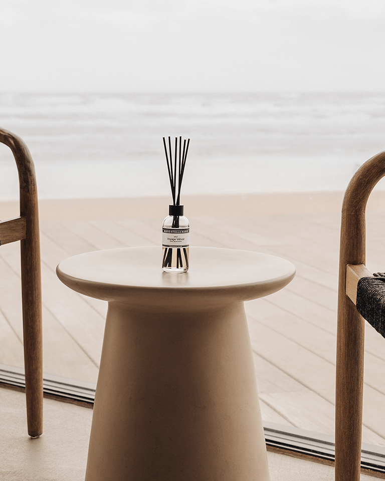 Marie-Stella-Maris Fragrance Sticks No.07 Voyage Vétiver 100 ml in a clear glass bottle with black reeds, placed on a round minimalist table between wooden chairs, with a soft, neutral interior and outdoor view in the background.